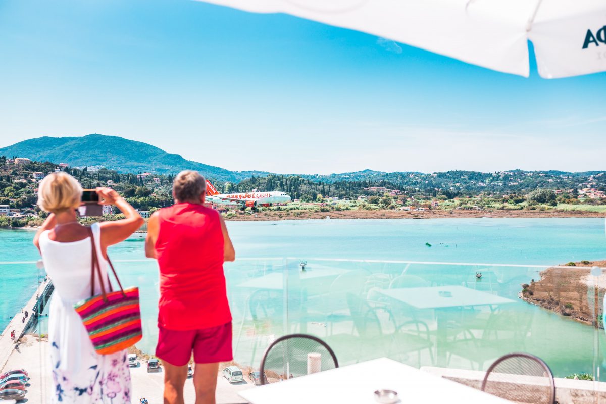 An airplane departing from Corfu, Greece on a sunny summer day. Two people are photographing the seaside landscape as the plane takes off.