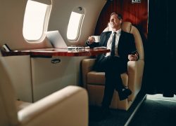 Senior businessman in suit flying in private jet and listening to music via wireless earphones.